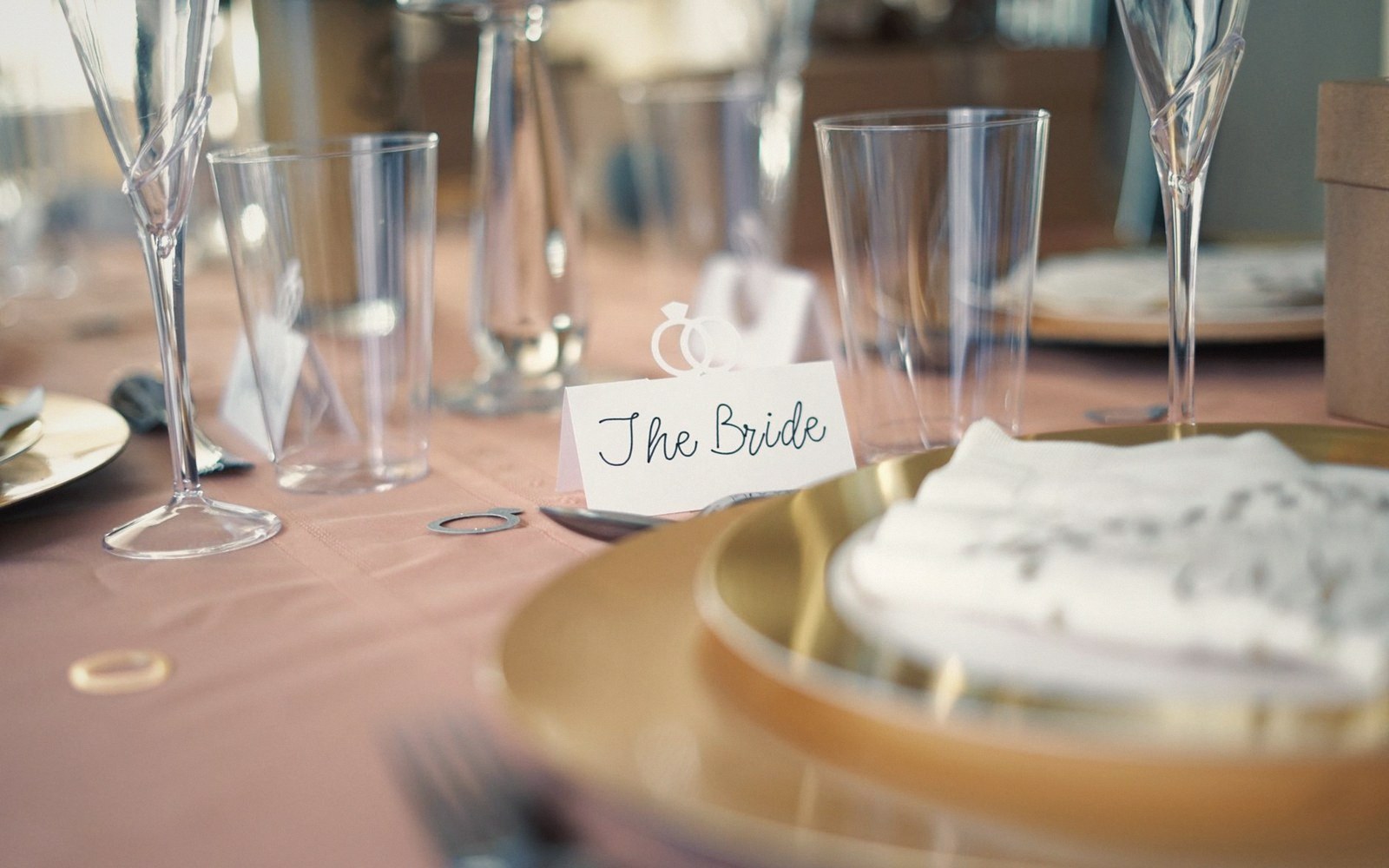 A place setting with a "The Bride" name card beside glassware and a gold charger plate