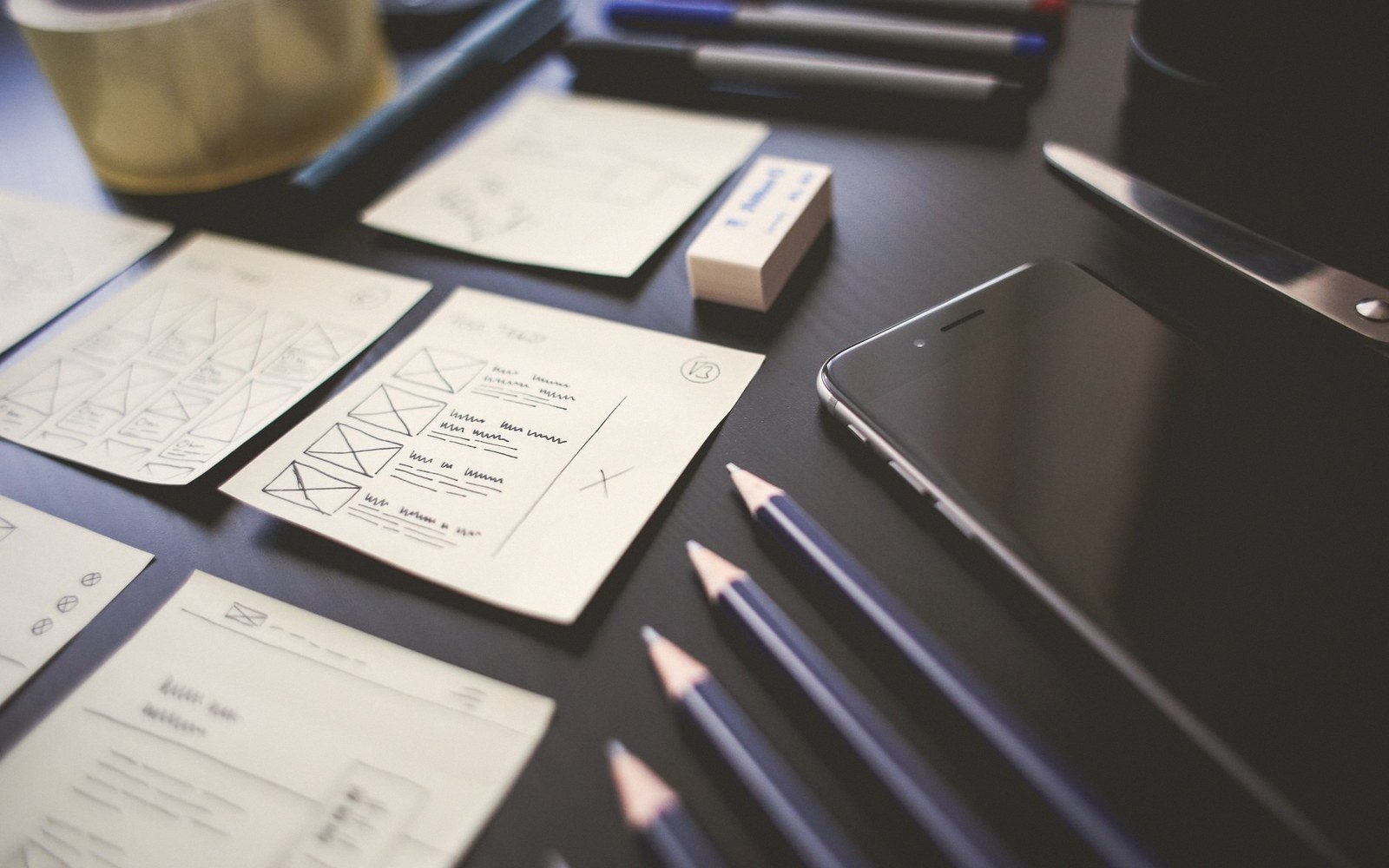Hand-drawn wireframe sketches on notepads beside pencils, a smartphone, and scissors on a desk